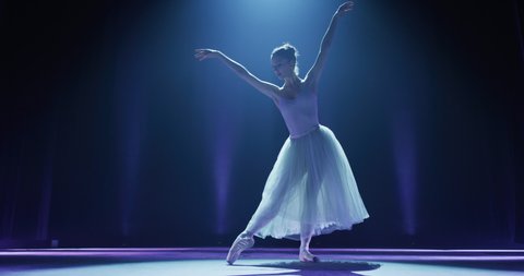 Cinematic shot of an young graceful classical ballet female dancer in white tutu is performing a choreography on classic theatre stage with spotlights before start of a show.