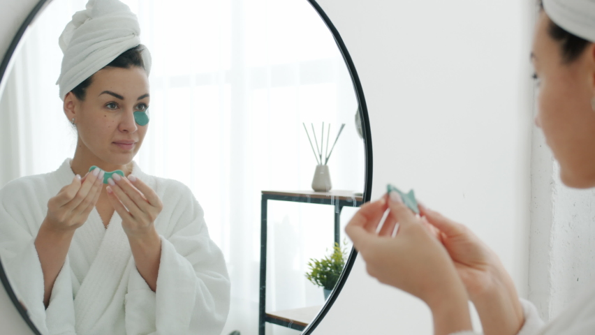 Beautiful young woman is applying eye patches caring for skin looking at mirror in bathroom and smiling. Beauty activities and people concept.