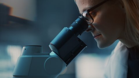 Close Up Portrait of a Medical Research Scientist Conducting DNA Experiments Under a Microscope in a Biological Applied Science Laboratory. Beautiful Female Lab Engineer in White Coat.