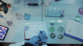 Above the Head Camera Footage of Medical Research Scientists in Blue Rubber Gloves Working Behind Tables in a Modern Laboratory. Lab Assistants Compare Samples, Use Tablet and Microscope. - Powered by Shutterstock - Get 15% off with code: PIKWIZARD15