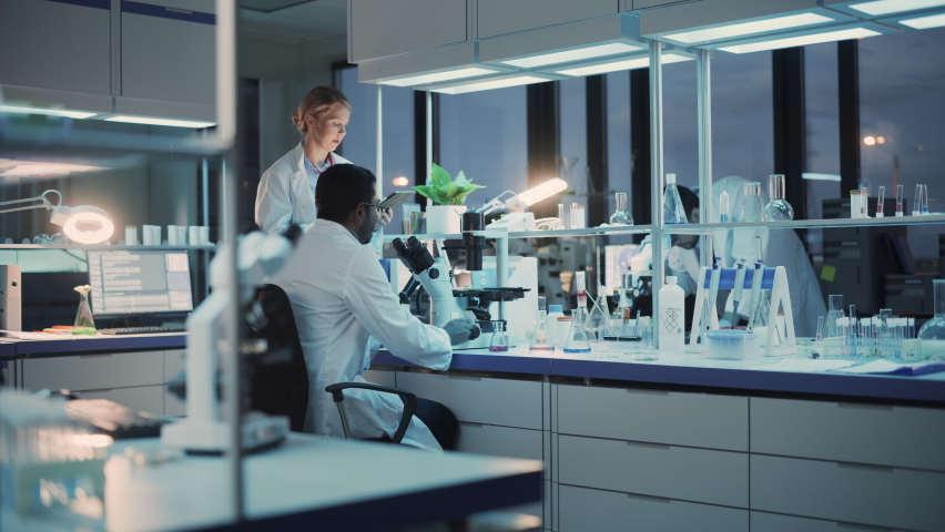Female and Male Medical Research Scientists Have a Conversation While Conducting Experiments Under a Microscope, Writing Analysis Results on a Tablet Computer. Modern Biological Science Laboratory. | Shutterstock HD Video #1061506546
