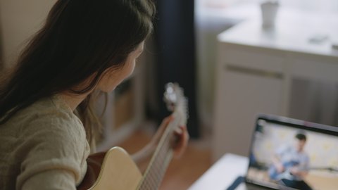 Stay home, solo activity Pretty girl watching online lesson and practising music. Young asian woman playing guitar at home. Aspiring musician learning to play musical instrument using laptop.