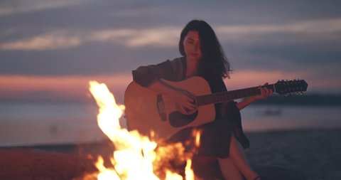 Lonely girl sitting by bonfire on the beach near forest playing acoustic guitar. Solo activity and social distancing.Spend free time in nature. Woman enjoying her loneliness. Summer vacation concept