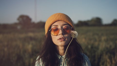 A beautiful young woman in stylish sunglasses stand in a field and blow a dandelion. A smiling hipster girl wearing trendy outfit enjoying summer sun in countryside. Concept of youth and creativity
