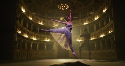 Cinematic shot of young couple of classical ballet dancers is performing a choreography on classic theatre stage with spotlights before start of a show.