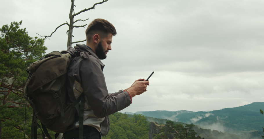 A man is standing on top of a mountain and typing a message on his smartphone. He has a large backpack on his shoulders. Hiking in the mountains. Top of the mountain. 4K DCI Royalty-Free Stock Footage #1061509591