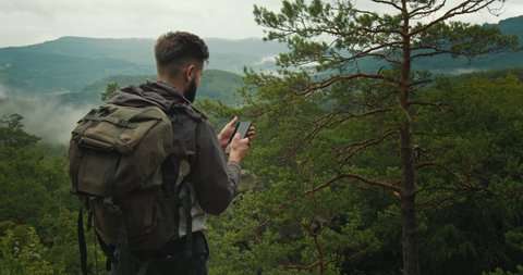 A man is standing on top of a mountain and typing a message on his smartphone. He has a large backpack on his shoulders. Hiking in the mountains. Top of the mountain. 4K DCI