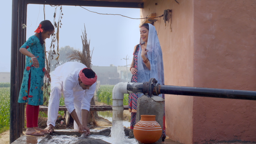 Happy Indian farmer and his small family playing with freshwater near a tubewell. Beautiful landscape view - Village couple and their cute daughter enjoying and laughing together outside a cottage Royalty-Free Stock Footage #1061510935