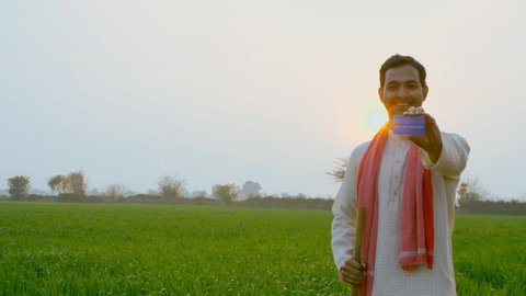 Pan shot of a confident Indian farmer happily showing his new debit or credit card. Medium shot of a self-sufficient villager in kurta-pajama smiling while holding his first Atm card - agricultural