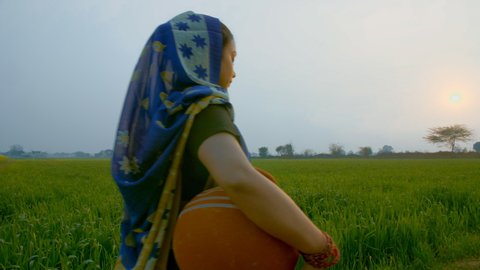 Village scene - Beautiful married woman walking through a field with a clay pot. Long shot of an Indian woman in a traditional saree carrying a pot of drinking water from a tubewell for her housework
