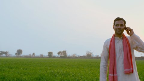 A smiling Indian farmer talking on mobile while standing near a green farmland. Medium shot of a happy villager wearing kurta-pajama, busy over a phone call - modern technology