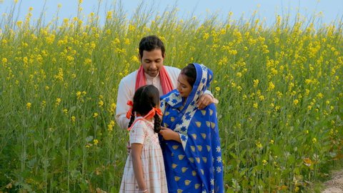 Cheerful Indian village family standing in front of their mustard (Sarso) field. Medium shot of a married couple with their little daughter, smiling and posing towards the camera - village scene