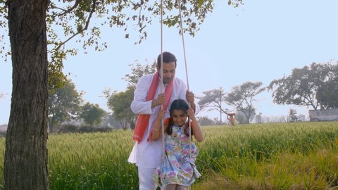 Cute little girl happily playing with her father on a tree swing during leisure time. Indian farmer spending quality time with his daughter in their agricultural field - happy family, parenting