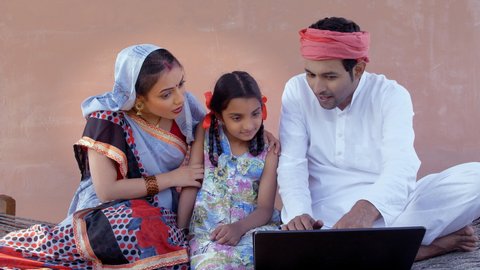Educated Indian farmer teaching the usage of a laptop to his wife and daughter. Happy nuclear village family learning new age technology together - working on laptop, village scene