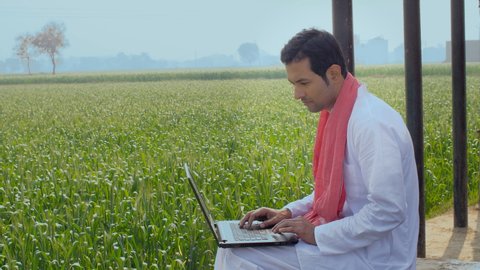 Educated Indian farmer learning new things on a laptop - technology concept. Young villager using laptop and internet while sitting on a Charpai - Indian agricultural field in the background