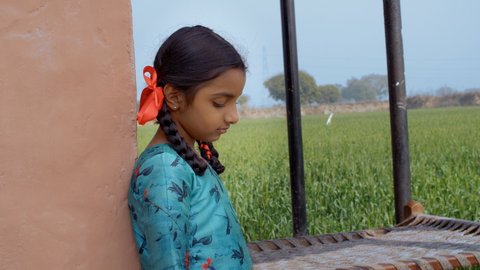 An upset small village girl stares out towards a field - the loneliness of a child concept. A sad child standing alone near green agricultural farmland