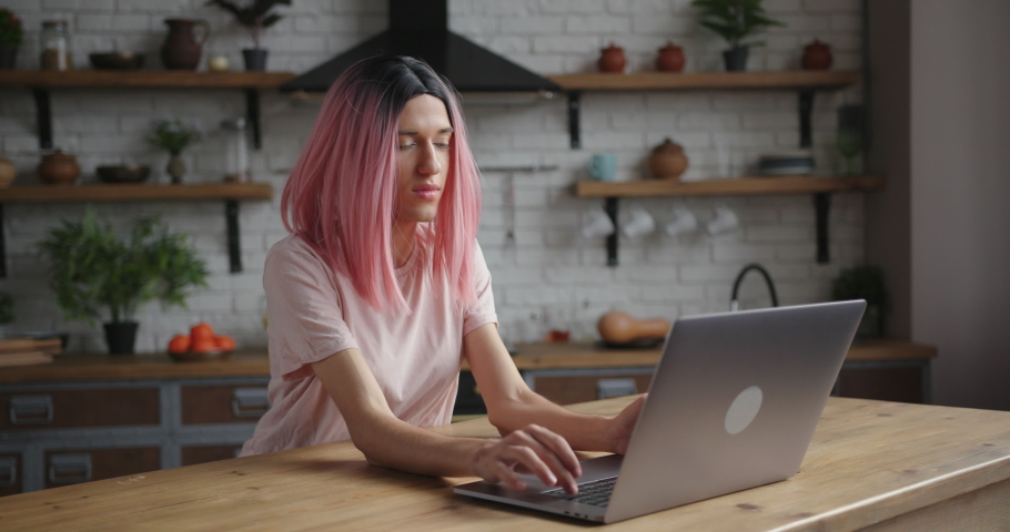Happy transwoman with pink wig has videocall via contemporary laptop sitting at wooden table in decorated kitchen at home