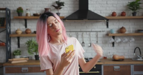 Cheerful young trans-woman with stylish pink wig surfs internet with contemporary cellphone standing in decorated kitchen closeup