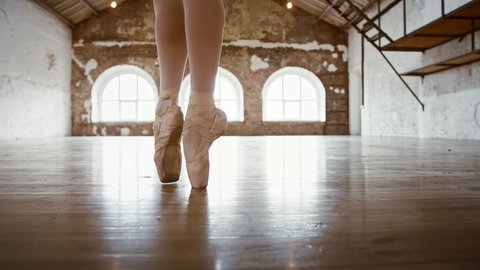 Legs of unknown ballerina in white nylon tights and pointe shoes spinning while dancing at a spacious loft with large windows letting sunlight in. Choreography and ballet. Close up, slow motion