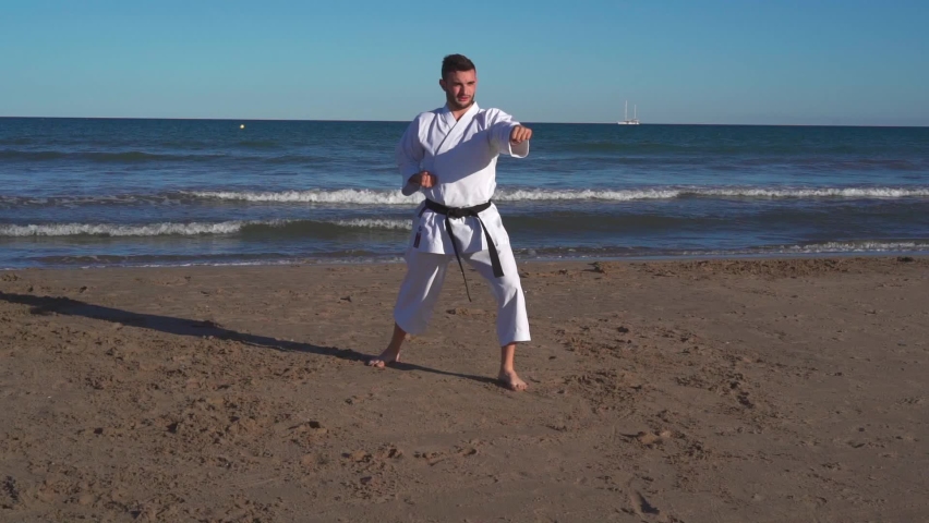 Karate fighter practicing kata on the beach at sunset and doing jumping, balancing and defense techniques in the open air. Wado ryu style. Truck right camera movement recorded in slow motion. kata