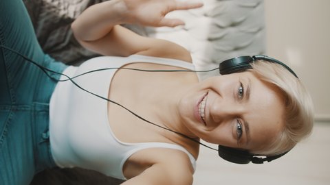 Vertical Shot Of Caucasian Girl In White Tank Top And Headphones Smiling, Waving And Blowing Kisses At Camera. - medium shot