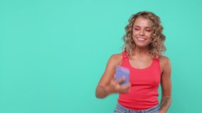 Excited young woman 20s in pink tank top isolated on blue turquoise background studio. People lifestyle concept. Talking on mobile phone making video call waving greeting with hand showing thumb up