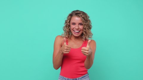 Smiling laughing young woman 20s years old in pink tank top posing isolated on blue turquoise background studio. People lifestyle concept. Pointing index fingers on camera blinking showing thumbs up