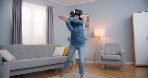 Good-looking Caucasian female in vr glasses training in virtual reality while being at home. Pretty young woman doing sports, playing tennis video game in living room. Technology concept.