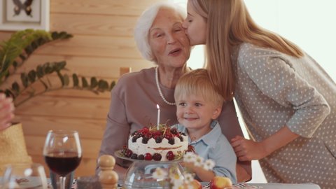 Cheerful senior woman sitting with cute little grandson at holiday dinner table while girl bringing birthday cake and kissing her cheek, kid blowing candle and family members clapping hands