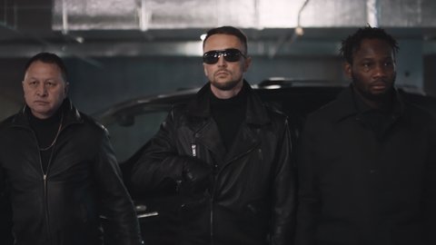 Slowmo walk of three multi ethnic criminals down underground parking lot leader in sunglasses getting gun out of leather jacket with black car in background
