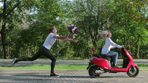 A guy with a bouquet of flowers runs after a girl on an electric moped. The guy wants to give flowers to the girl.
