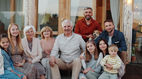 Happy multigenerational Caucasian family and dog looking together at camera, smiling and waving while sitting outdoors on wooden terrace of vacation house decorated with lights