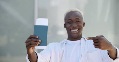 Outdoors portrait young african american guy man foreign student joyfully shows holding ticket and passport documents for boarding plane train, feeling happiness from travel trip, adventure concept
