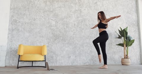 Young girl woman athlete with long hair in black sportswear stretches at home raises her legs in turn stretches muscles prepares for dance rehearsal workout at home in room against background of walls