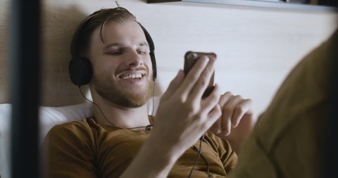 Portrait of young positive man in headphones using video chat. Cheerful handsome Caucasian guy messaging online lying on bed. Smiling man waving and talking. Cinema 4k ProRes HQ.