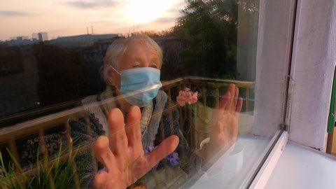 The Covid-19, health, safety and pandemic concept - senior old lonely woman wearing protective medical mask sitting near the window at home for protection from virus