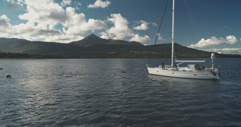 Amazing seascape of ocean bay with yacht aerial. Green mountain island with forest at mist haze. Epic sail boat at sea harbor of Brodick pier, Arran island, Scotland, Europe. Cinematic summer cruise