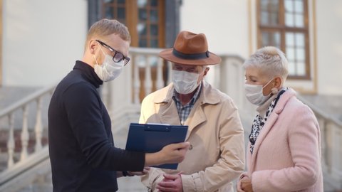 Male real estate agent and senior couple in safety mask discussing over documents outdoors. Young broker showing house plan to aged man and woman clients wearing protective mask