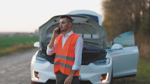 Man calling car assistance services because his electric car is broken. Concept road accident. Help repair. Man in a safety vest talking on cell phone.