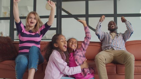 Excited joyful multiethnic family with two lovely elementary age mixed race daughters sitting on couch, cheering favorite team and emotionally celebrating goal while watching sport on tv at home.