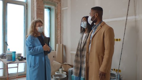 Female designer in safety mask showing house renovation process to young african couple. Real estate agent and afro-american family wearing protective mask observing apartment on sale