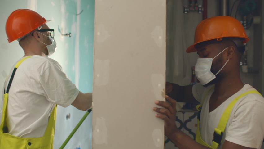 Diverse migrant workers in safety mask renovating customer apartment. Side view of caucasian builder painting walls and african colleague installing plumbing at construction site