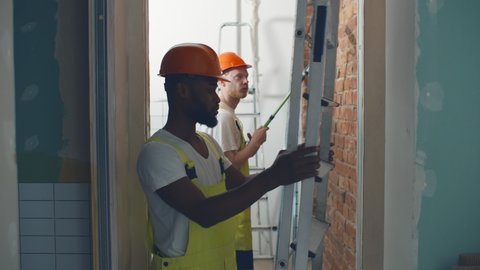 African worker man carrying aluminum ladder doing apartment renovation. Black foreman in hardhat and overall walking with stepladder in redecorated new house