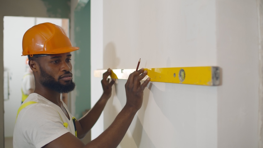African male worker checking level of the wall with the bubble level tool. Portrait of afro-american builder using spirit level inspecting apartment renovation