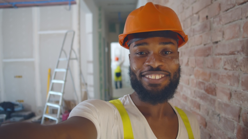 Pov shot of african builder in safety helmet taking selfie over construction site background. Smiling afro immigrant worker renovating apartment and filming video for internet blog Royalty-Free Stock Footage #1061546968
