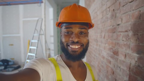 Pov shot of african builder in safety helmet taking selfie over construction site background. Smiling afro immigrant worker renovating apartment and filming video for internet blog