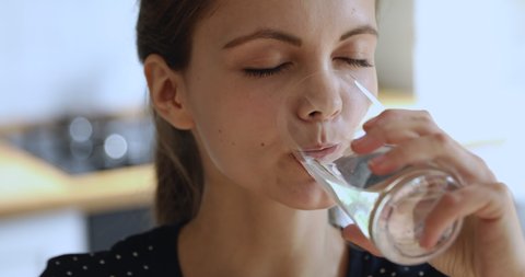 Close up head shot healthy happy positive young attractive woman making sips, drinking stilled pure mineral water from glass, normalizing bowel function, feeling good at home, daily healthcare habit.
