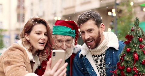 Close up portrait of happy Caucasian female and male standing with Santa Claus and taking selfie pictures on street. Joyful couple with Santa recording video for vlog. Christmas spirit concept