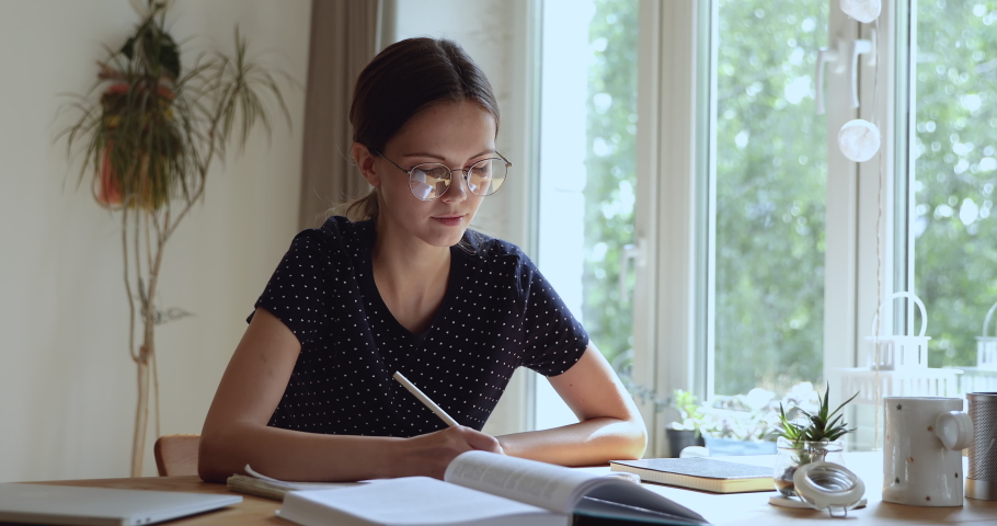 Smart young teen girl in glasses studying at home, reading books, making noted in paper copybook, preparing for examination or session, learning subject indoors, university higher education concept. Royalty-Free Stock Footage #1061553169