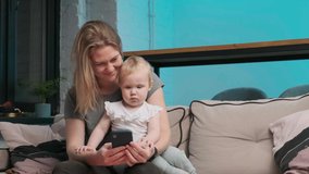 Young mother in a gray T-shirt orders food on phone, her daughter also looks at phone and they happy. child pokes at smartphone screen and nods while they sit on couch. great option for mockup.